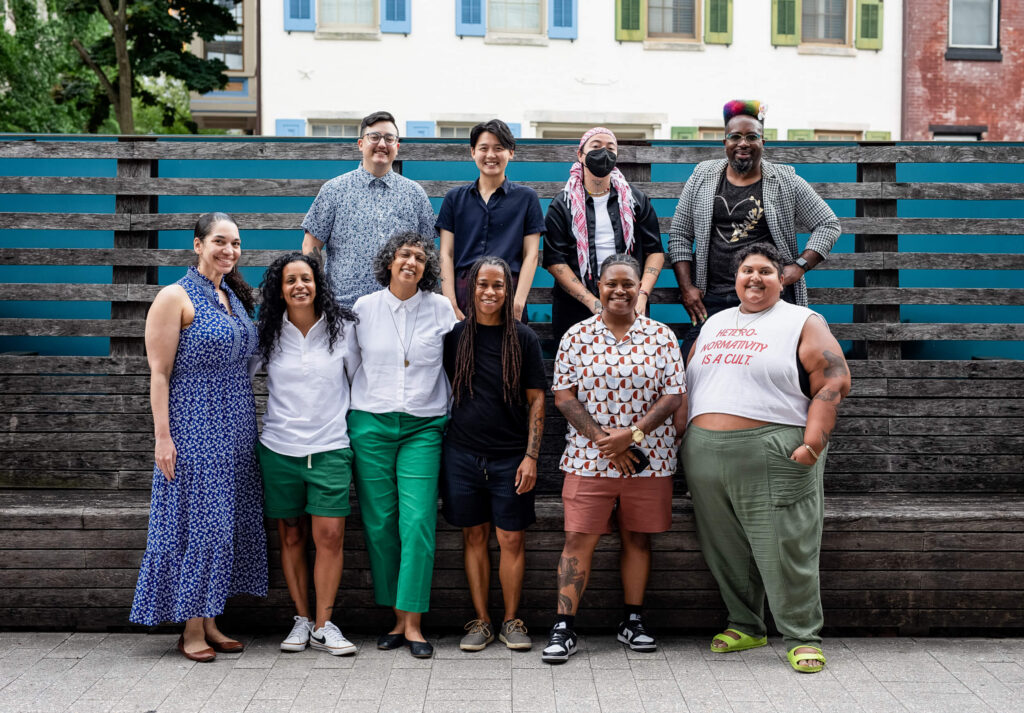 Ten NQTTCN members, including the Founding Director Erica Woodland, pose together at a regional gathering. They are outside, standing in front of a striped wooden relief.