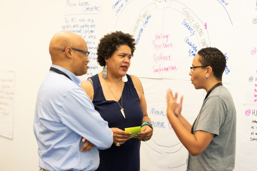 Cara Page, a co-architect of the Healing Justice framework and co-author of Healing Justice Lineages with Erica Woodland, talks with two other people in front of a wall covered with large sheets of paper displaying handwritten text and diagrams.